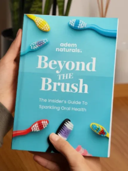 Person holding a book titled ‘Beyond the Brush: The Insider’s Guide to Sparkling Oral Health’ by Adem Naturals, featuring colorful toothbrushes on a blue cover.
