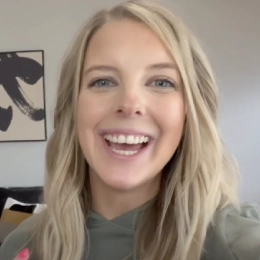 Smiling woman with blonde hair wearing a green hoodie, sitting indoors with a modern wall painting in the background.