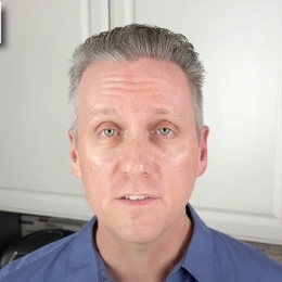 Man with short gray hair wearing a blue shirt, standing in a bright kitchen with white cabinets.